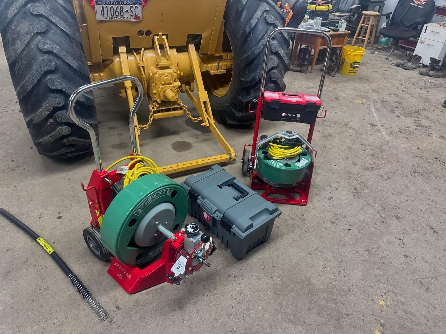 Two red industrial pipe cleaning machines, a gray plastic case, and a tractor in a workshop.