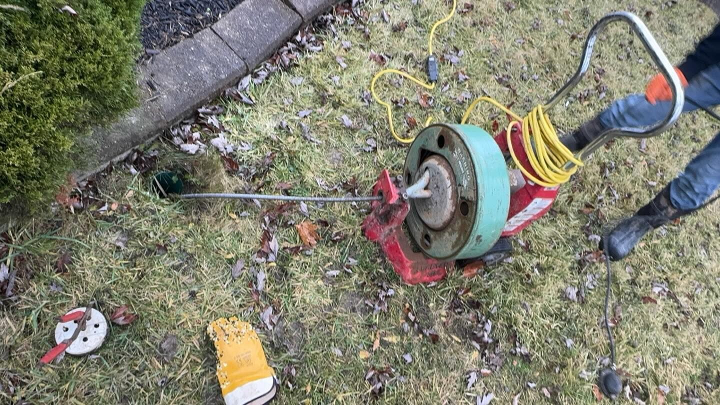 A power drain auger cable feeds into a ground pipe in a grassy yard, with a yellow glove lying nearby.