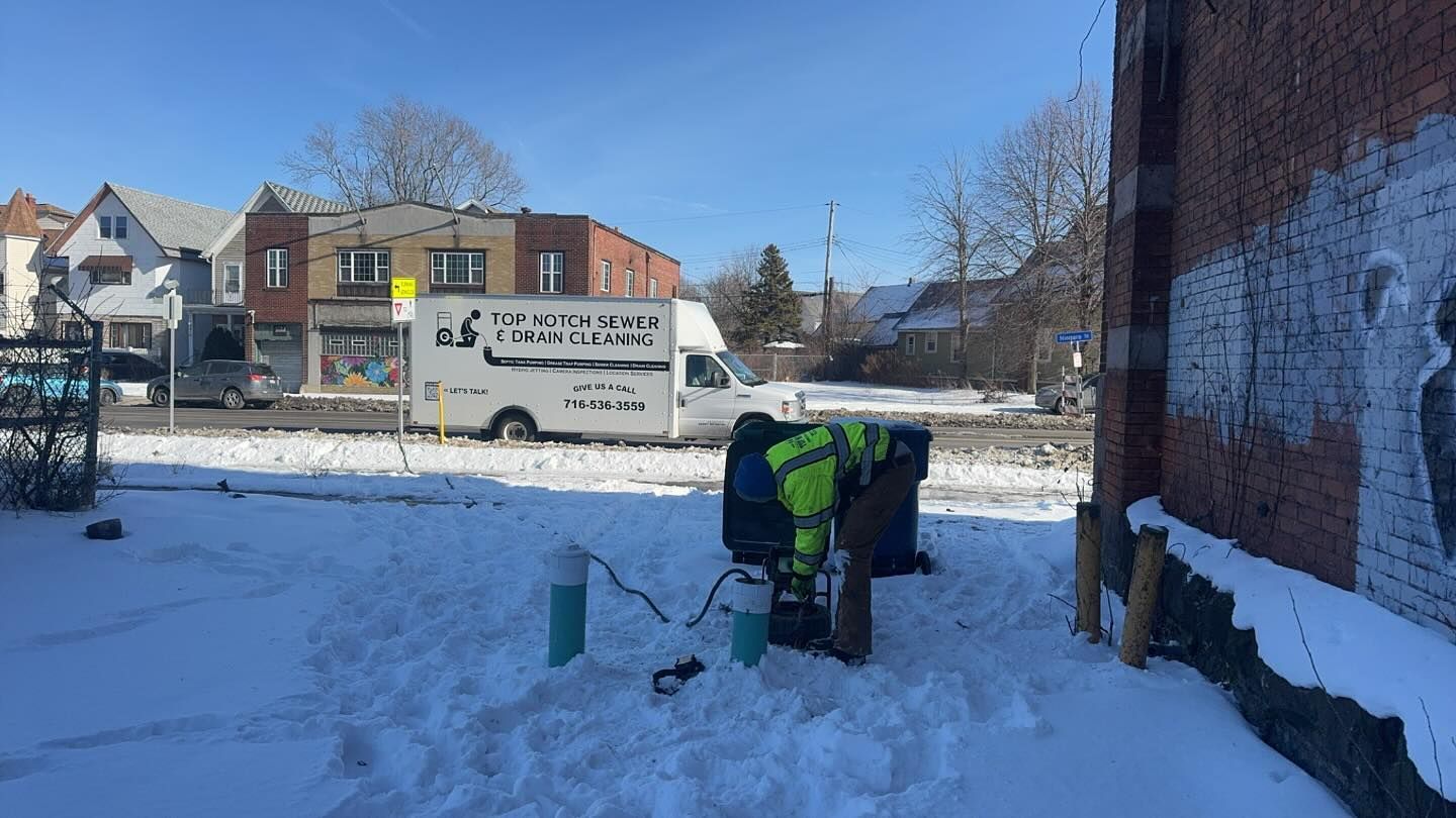 A worker in a high-visibility jacket tends to equipment in a snow-covered urban lot near a brick building.