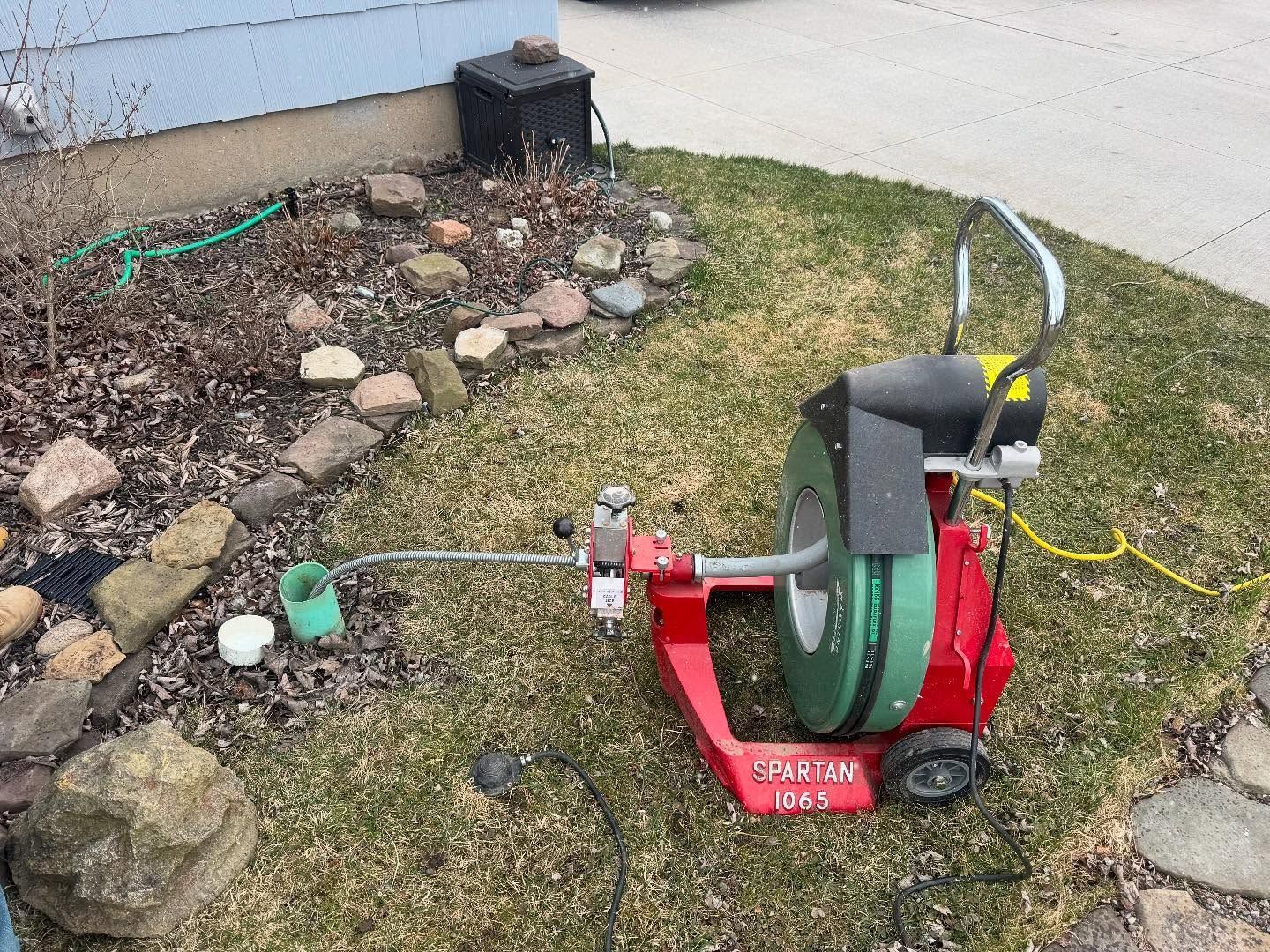 A red Spartan Tool drain cleaning machine is positioned on a lawn, with its metal cable extending into a ground pipe.