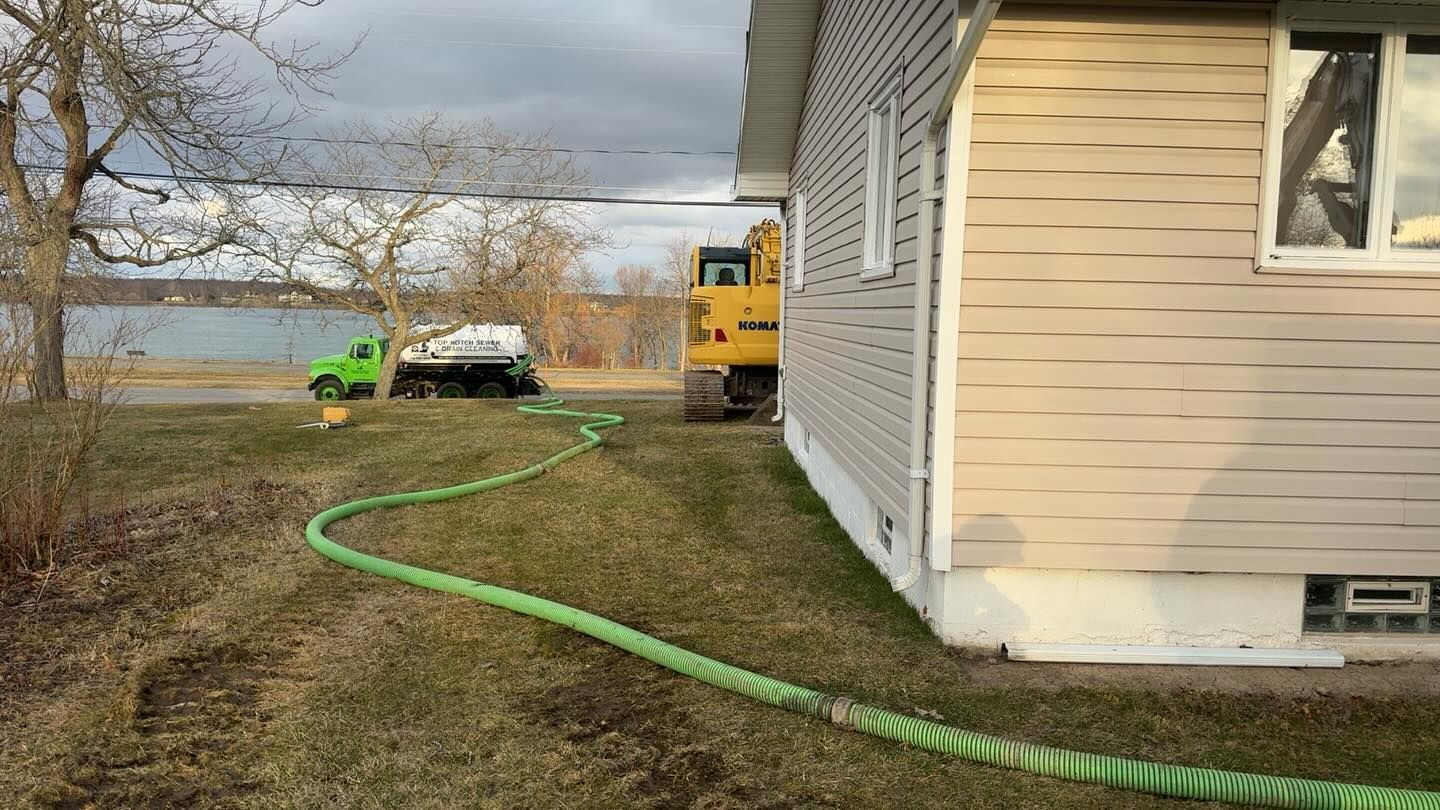 A long green hose leads from a lakefront property to a yellow excavator and white truck parked on a grass lawn.