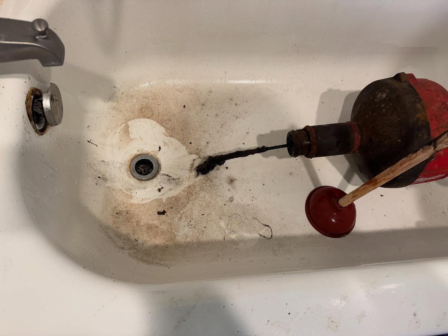 A drain snake removing a clump of hair and debris from a bathtub drain, with a red plunger resting nearby.