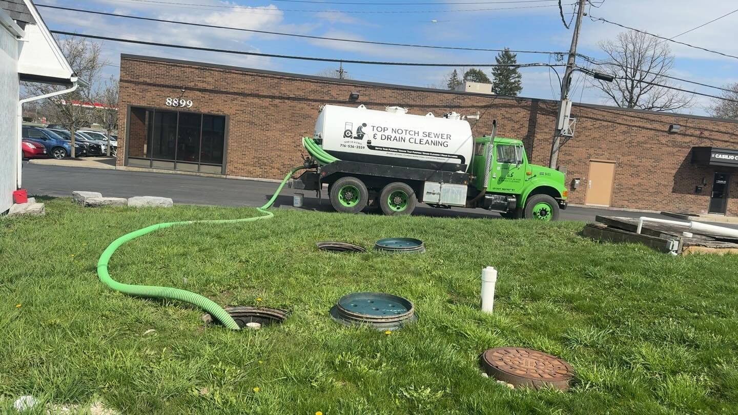 A bright green septic service truck parked on a street with a hose running to a buried tank in a grassy lawn.