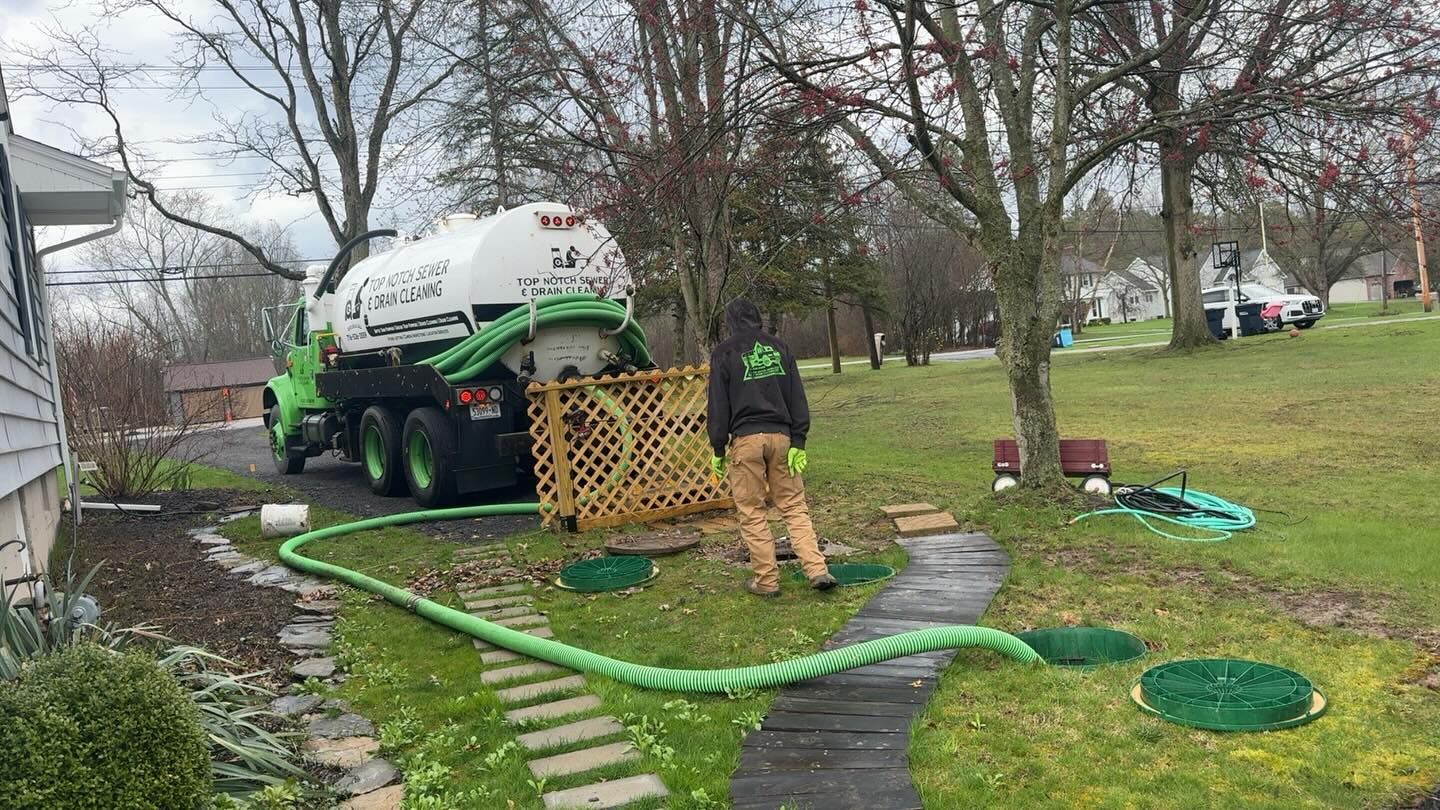 A technician in a uniform uses a hose connected to a large white septic service truck to pump out a residential tank.