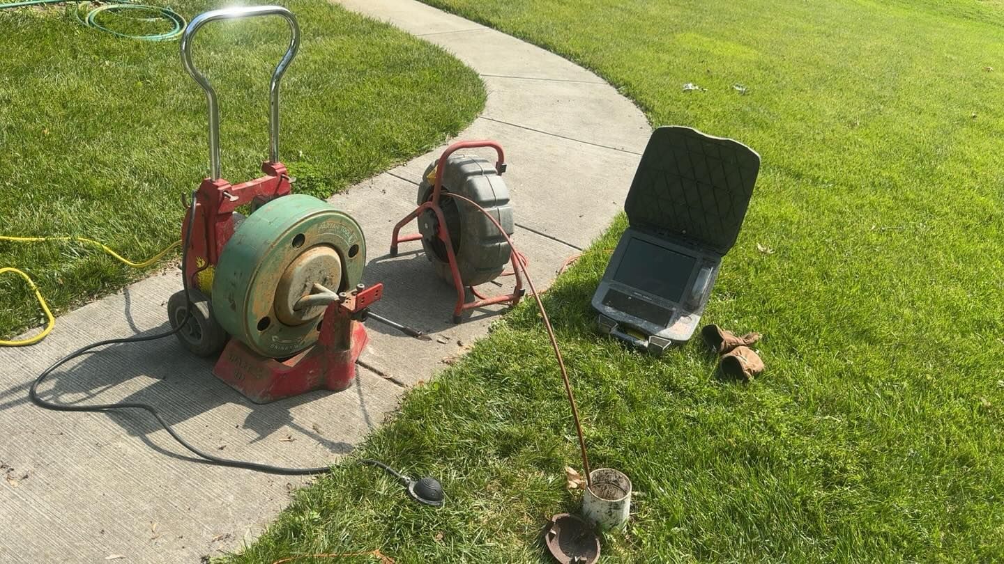 Sewer inspection camera equipment, including a red-and-green reel and monitor, sits on a sidewalk next to a grassy lawn.