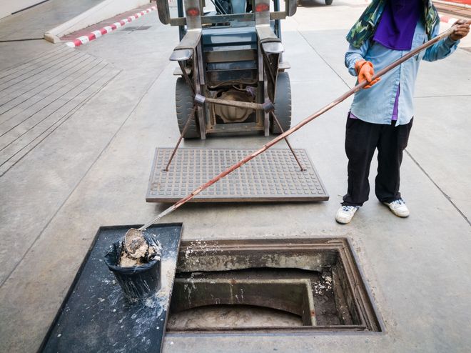 A worker uses a long-handled tool to retrieve a bucket of debris from an open storm drain near a forklift.