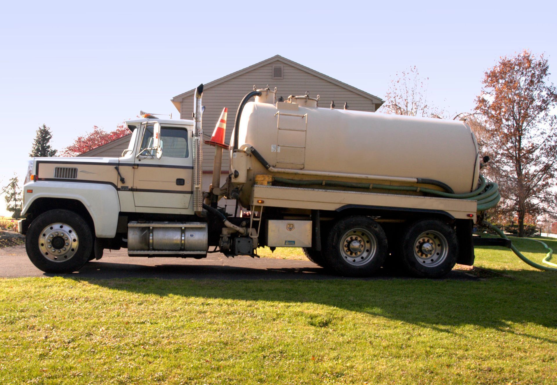 A light-tan tanker truck parked on a grass lawn in front of a house, with a green hose extending from the rear.
