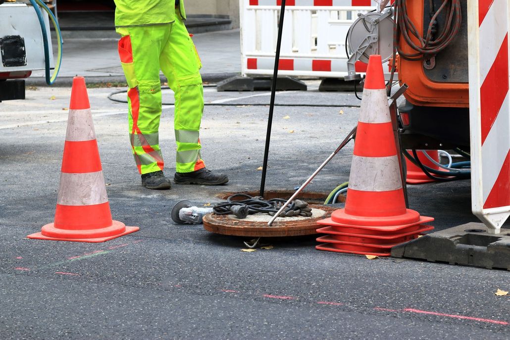 A worker in neon yellow safety gear stands near two traffic cones and an open manhole cover on a paved road.