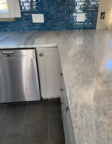 A kitchen counter with grey-and-white stone, a stainless steel dishwasher, white cabinets, and blue tiled backsplash.