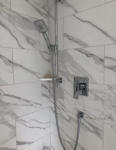A chrome shower head attached to a vertical sliding rail against white marble-patterned tile walls.
