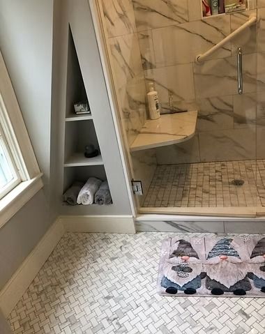 A marble-tiled bathroom with a triangular wall shelf, a glass-enclosed shower, and a decorative gnome-themed floor mat.
