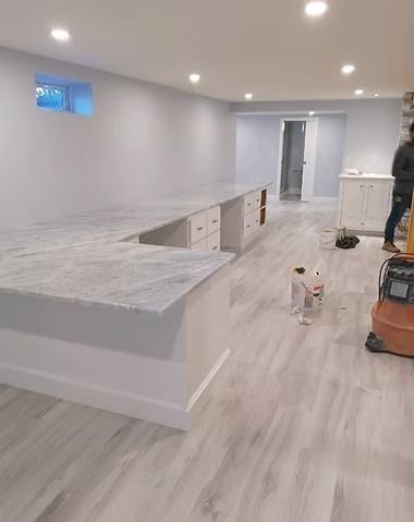 A modern basement renovation featuring a long, white U-shaped desk with a gray marble countertop and light wood-look flooring.