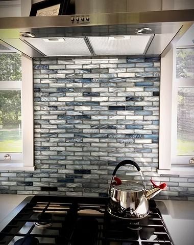A stainless steel tea kettle sits on a gas stove in front of a blue and white glass tile backsplash under a vent hood.