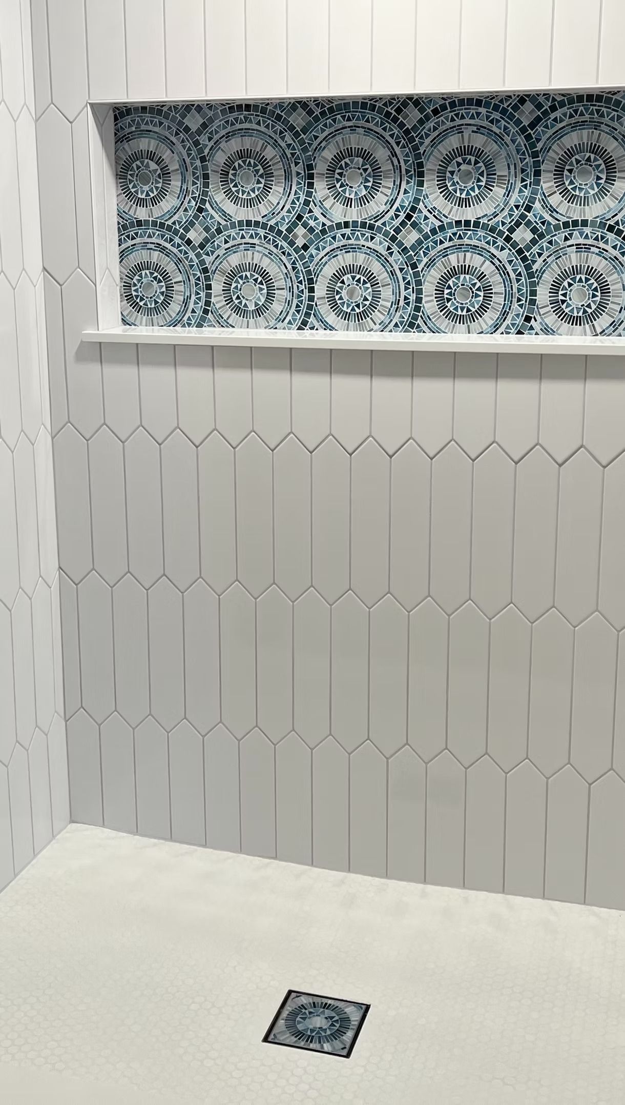 A shower stall with white picket-style wall tiles, a blue and white patterned accent niche, and a matching drain cover.