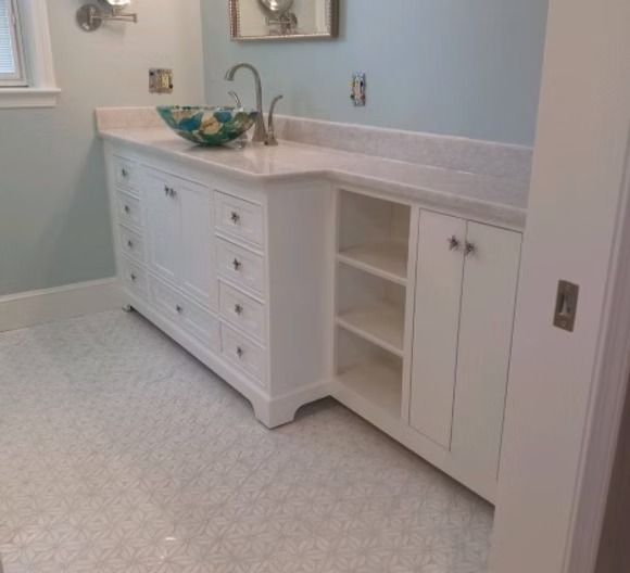 White bathroom vanity with a patterned glass vessel sink, marble countertop, open shelving, and cabinets on tiled floor.