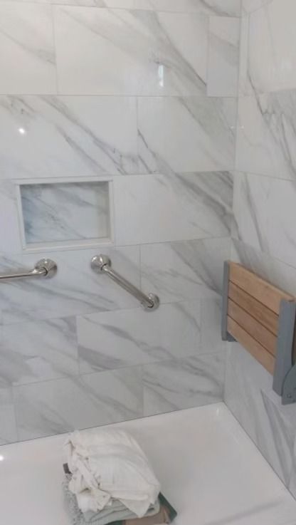 A marble-tiled shower interior featuring two chrome grab bars, a built-in shelf, and a wall-mounted wooden fold-down seat.