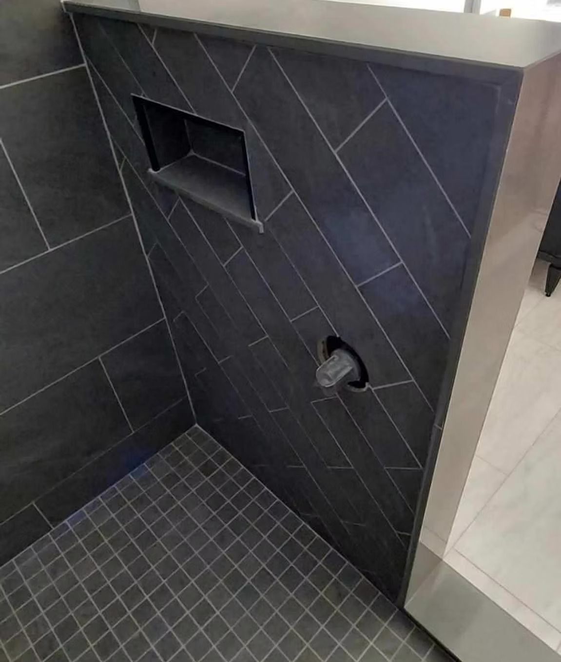 A modern shower wall featuring dark grey herringbone tiles, a built-in recessed shelf, and a protruding shower handle.