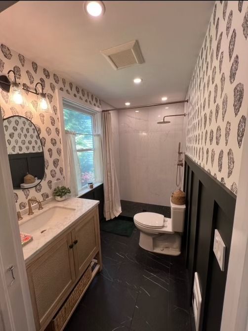A narrow bathroom with black-and-white wallpaper, black wainscoting, marble-look floors, a wooden vanity, and a shower area.