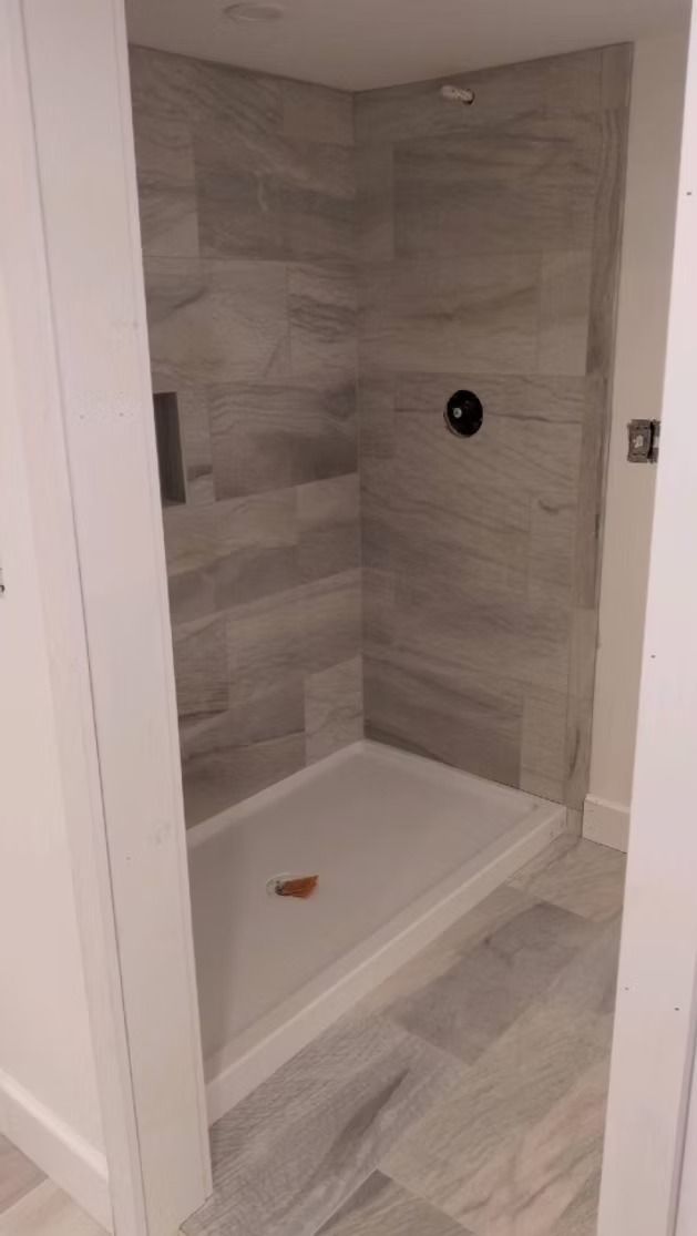 A walk-in shower with light grey wood-look wall and floor tiles, a white shower pan, and a black circular shower control.