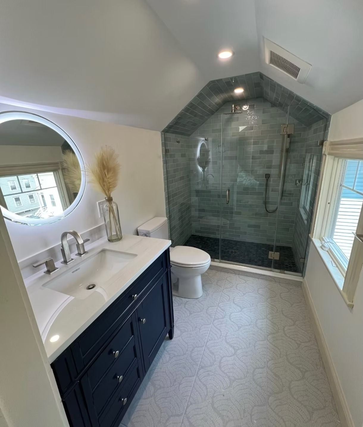 Narrow bathroom with double vanity, round mirror, white walls, and gray tiled shower alcove.