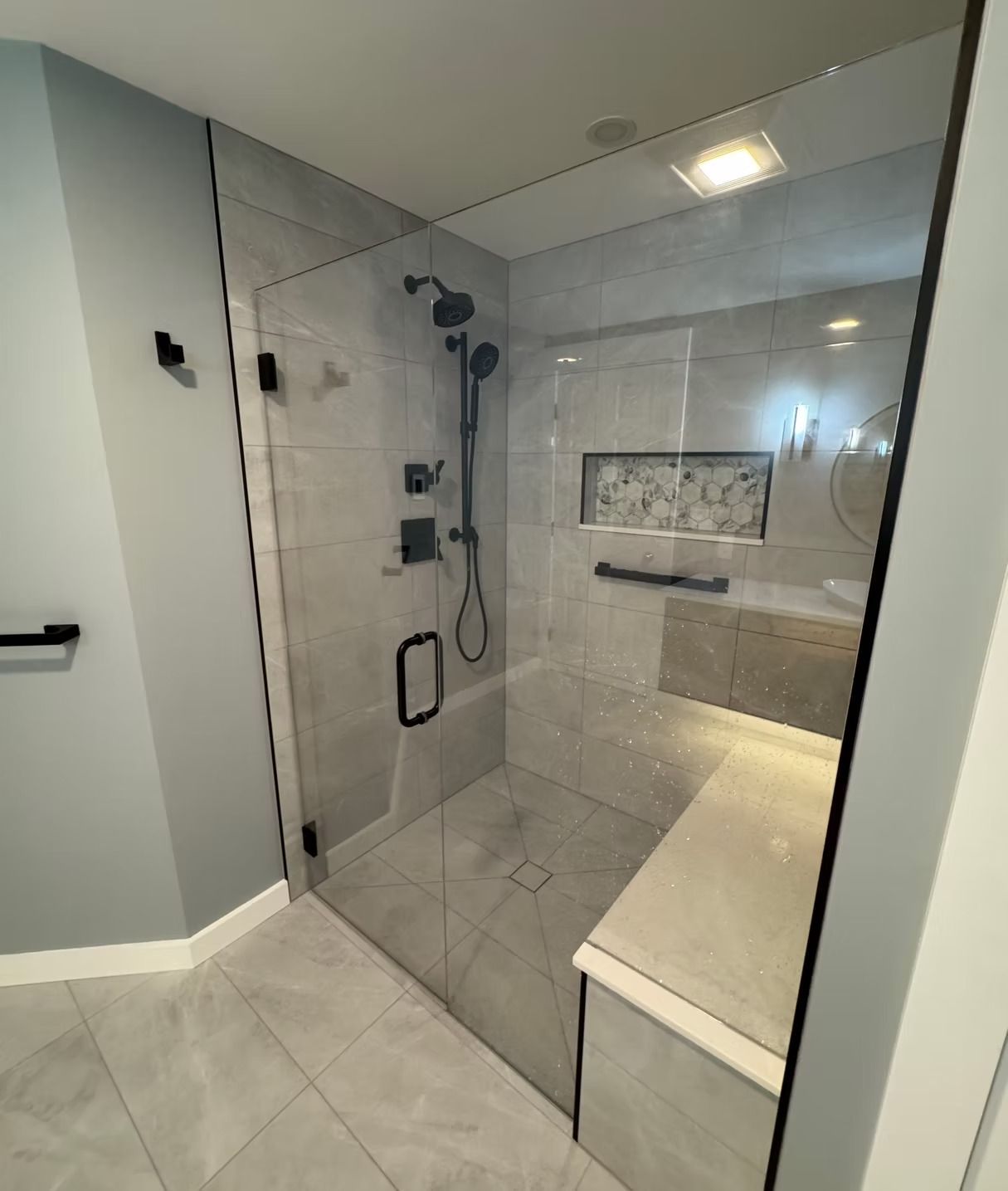 Modern walk-in shower with gray tile, a built-in bench, glass door, and black fixtures, including a handheld shower head.