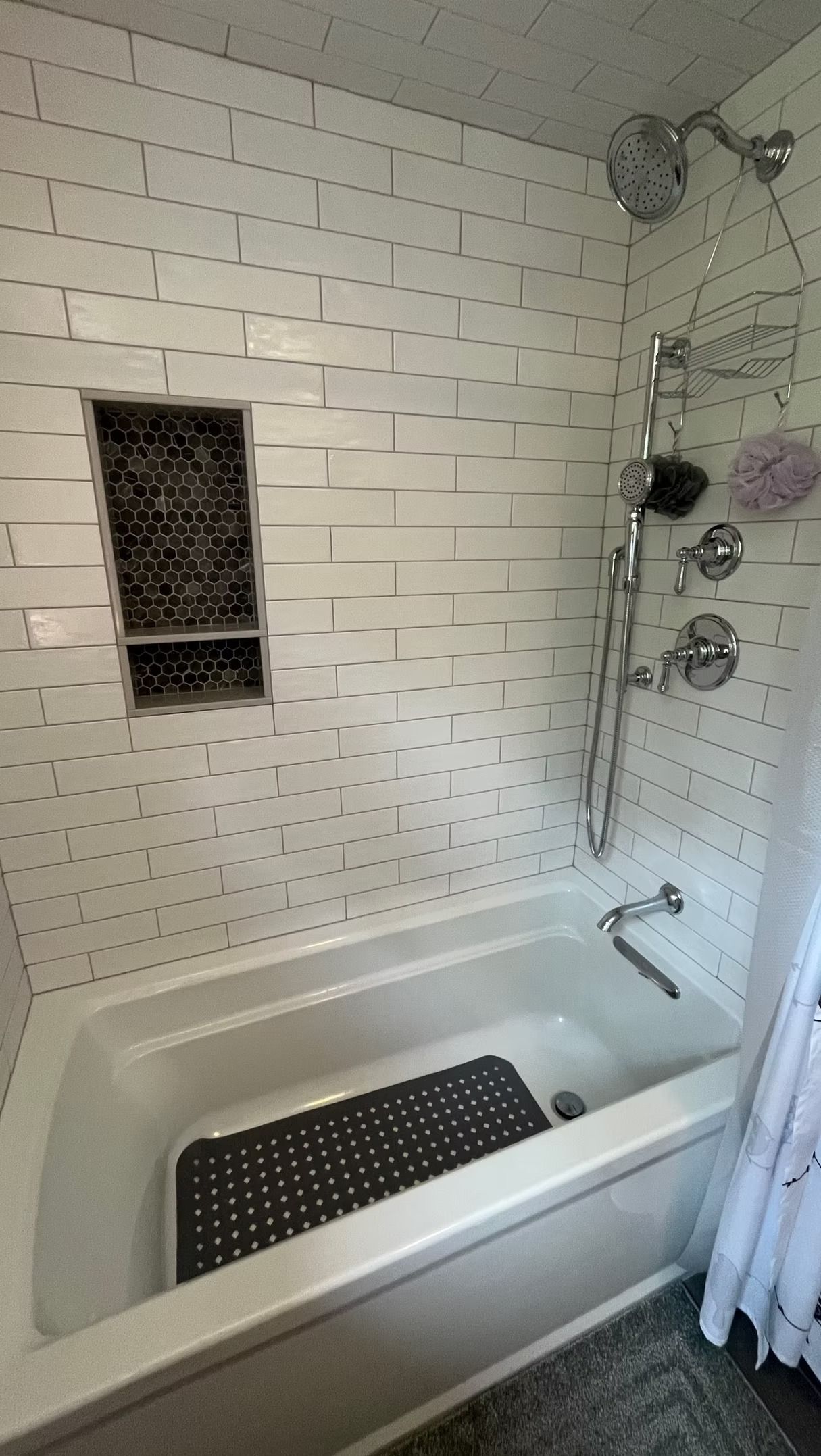 A white tiled bathtub and shower with a built-in storage niche, a showerhead, faucet, and a black non-slip mat inside.
