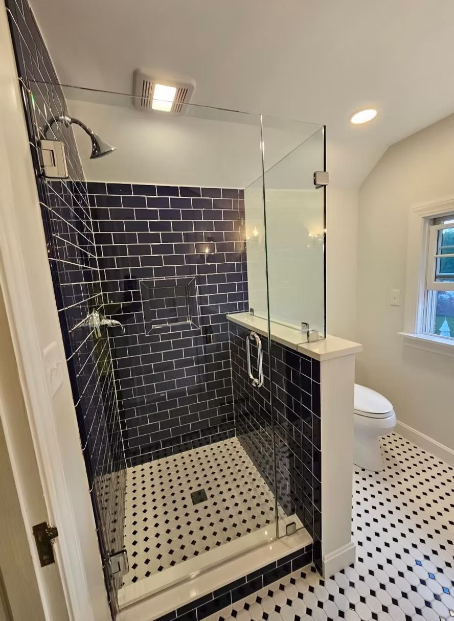 A walk-in shower with dark blue subway tiles and a black-and-white patterned tile floor, next to a white toilet.