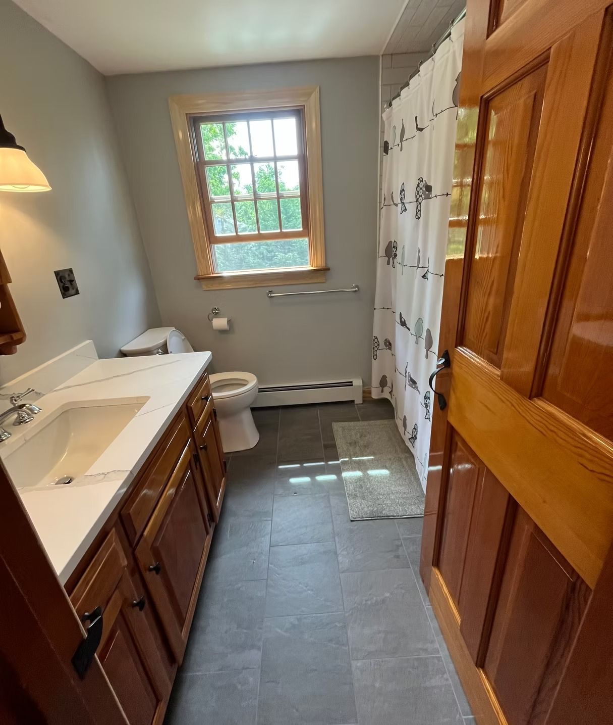 A bathroom with gray walls, a wooden vanity, a sink, a toilet, a shower with a patterned curtain, and dark tiled flooring.