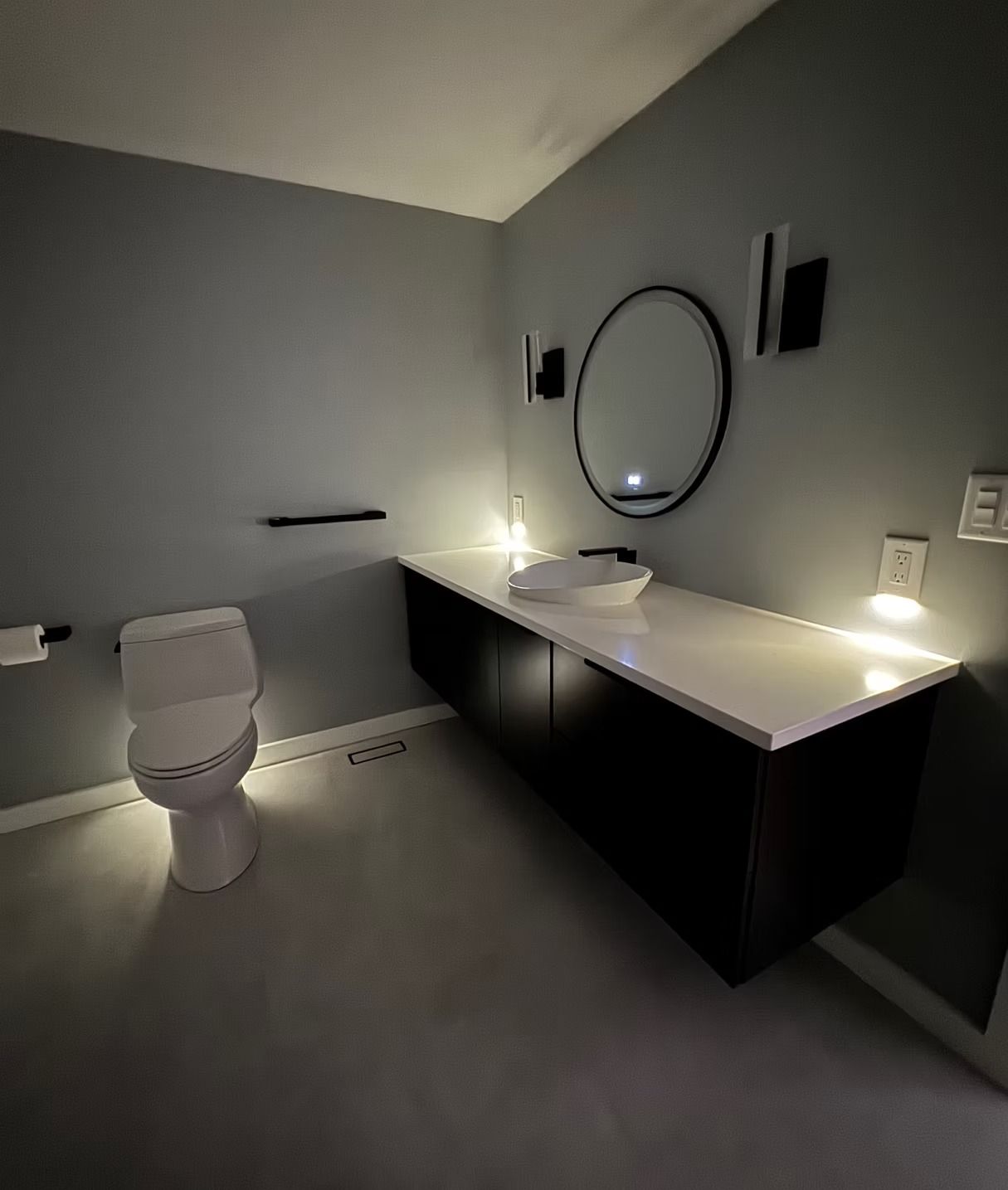 A modern, dimly lit bathroom with gray walls, a floating black vanity with a white countertop, and a circular mirror.