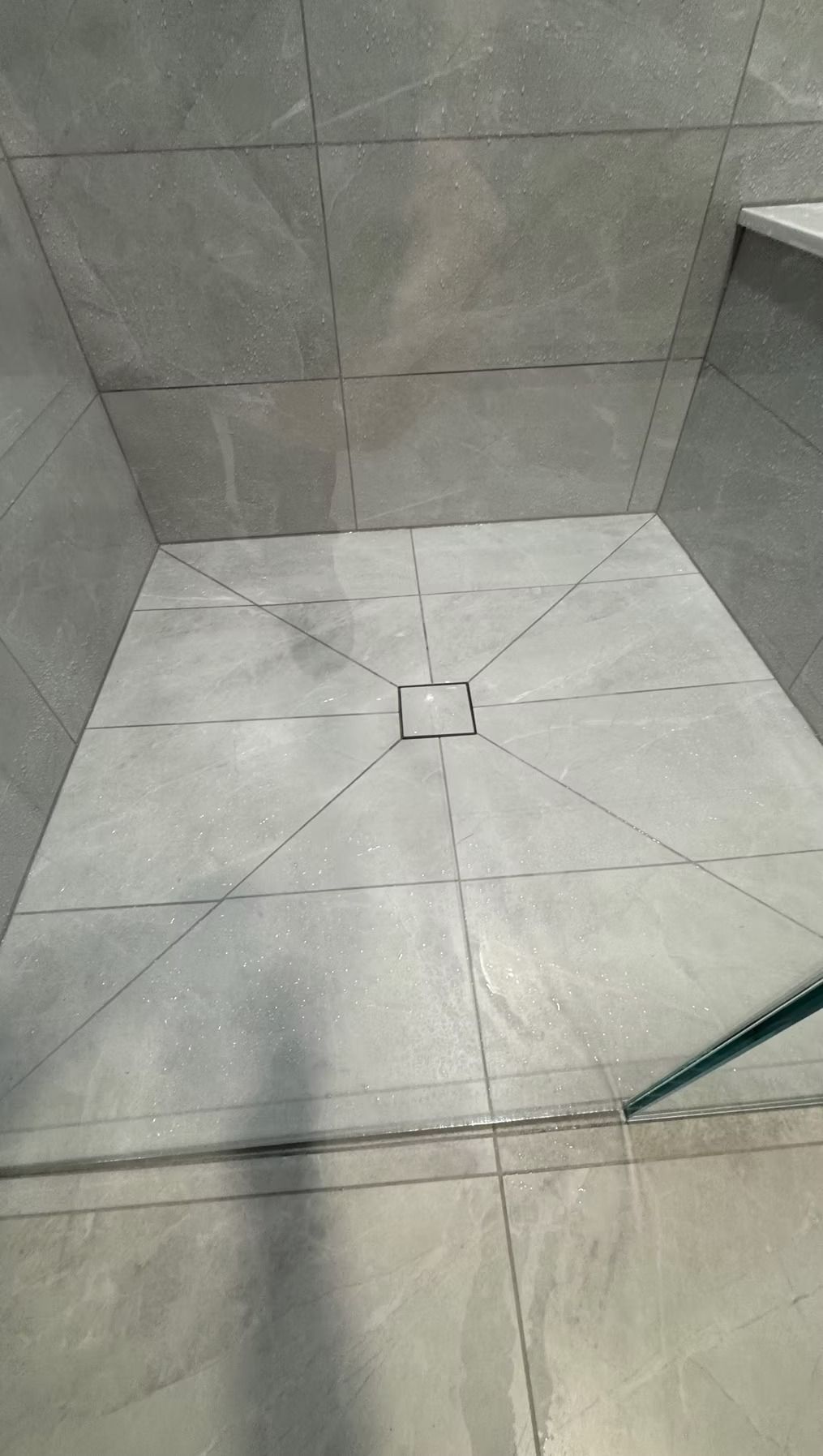 Cracked light-gray tiled shower floor with gray wall tiles and a glass edge visible on the right.