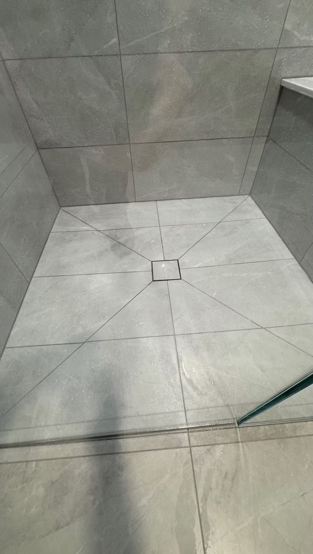 A square, gray-tiled walk-in shower floor with a central drain, featuring diagonal grout lines leading to the drain.
