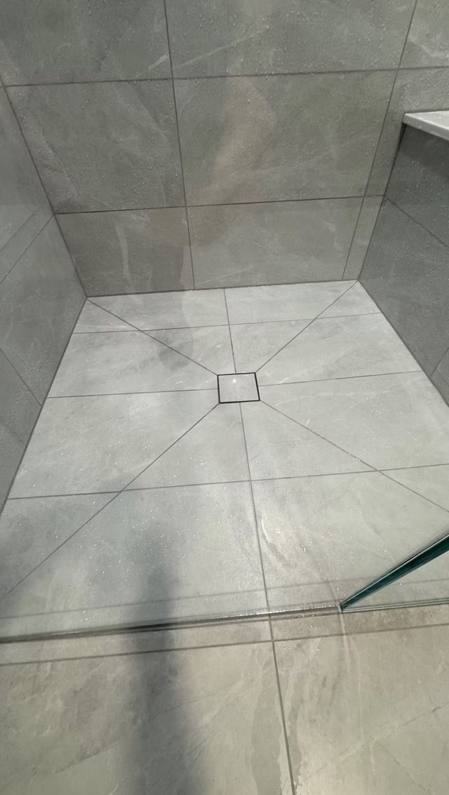 A square, gray-tiled walk-in shower floor with a central drain, featuring diagonal grout lines leading to the drain.