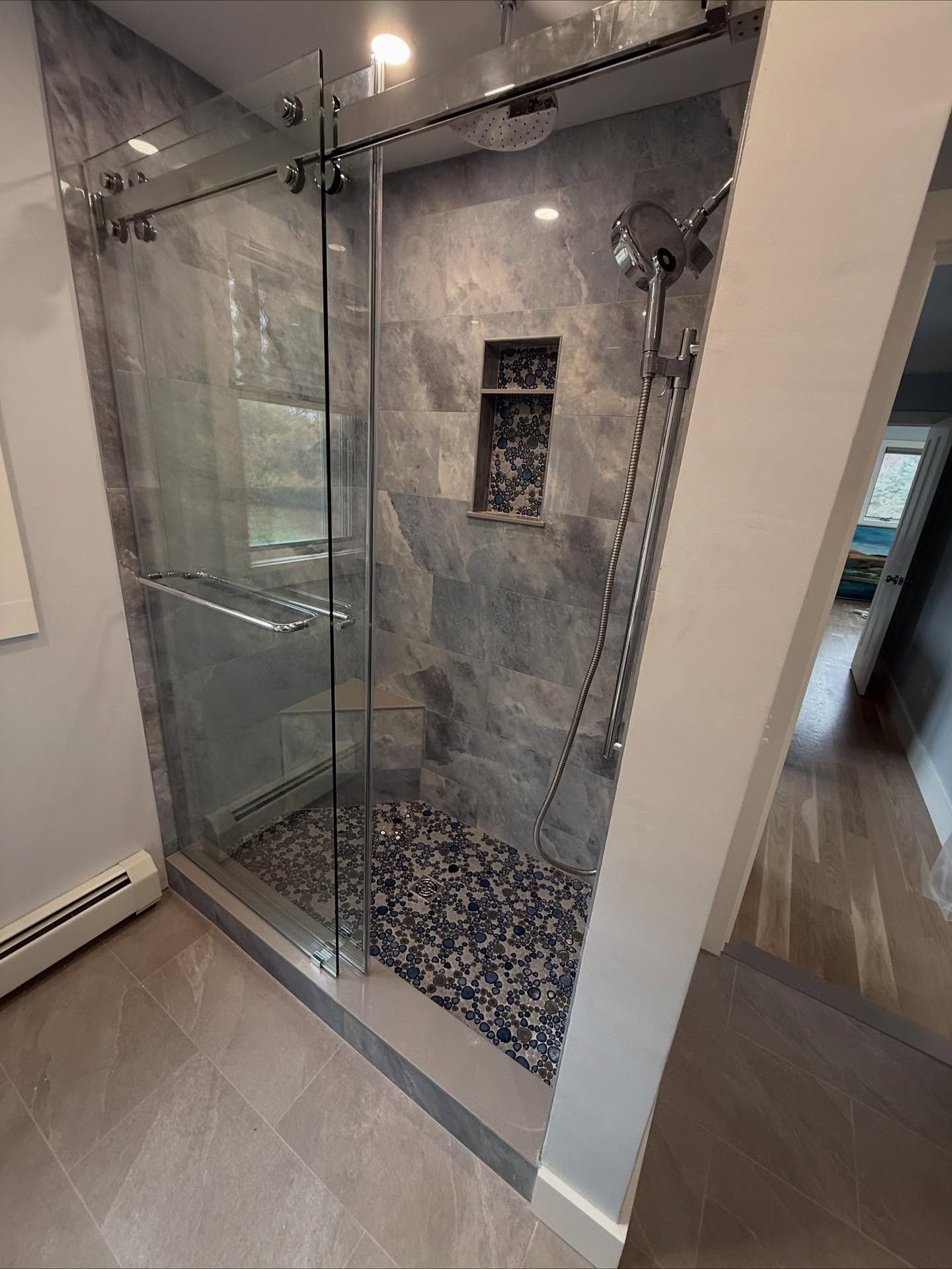 A modern glass-enclosed shower with gray tiled walls, a pebble floor, a small built-in shelf, and a chrome shower head.