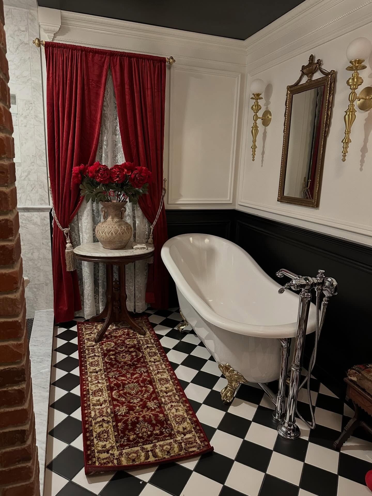 A bathroom with a clawfoot tub, checkered floor, red curtains, a vintage side table with roses, and gold wall sconces.