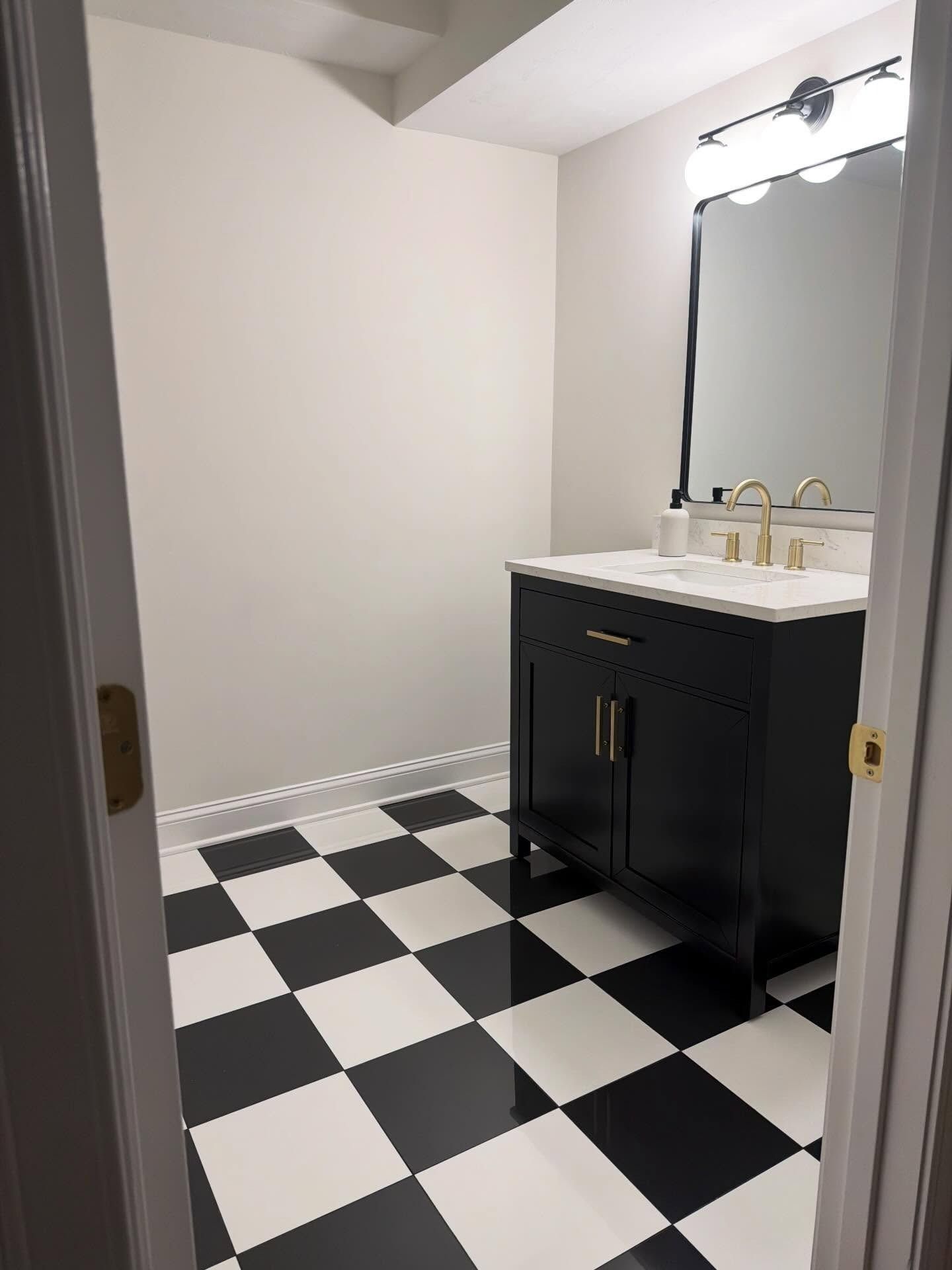 A modern bathroom with black and white checkered floor tiles, a black vanity cabinet with gold hardware, and a mirror.