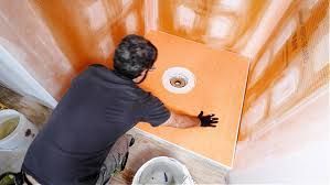 A person installing a waterproof shower pan liner in a tiled corner, pressing it down onto the subfloor around the drain.