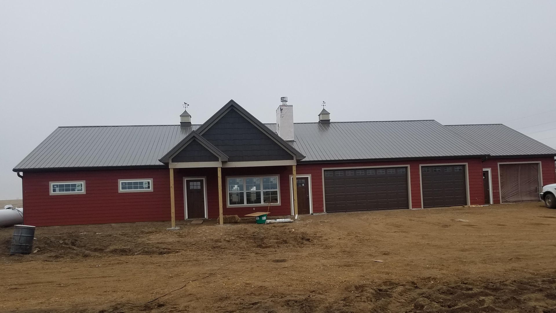 A large red house with a black roof is sitting in the middle of a dirt field.