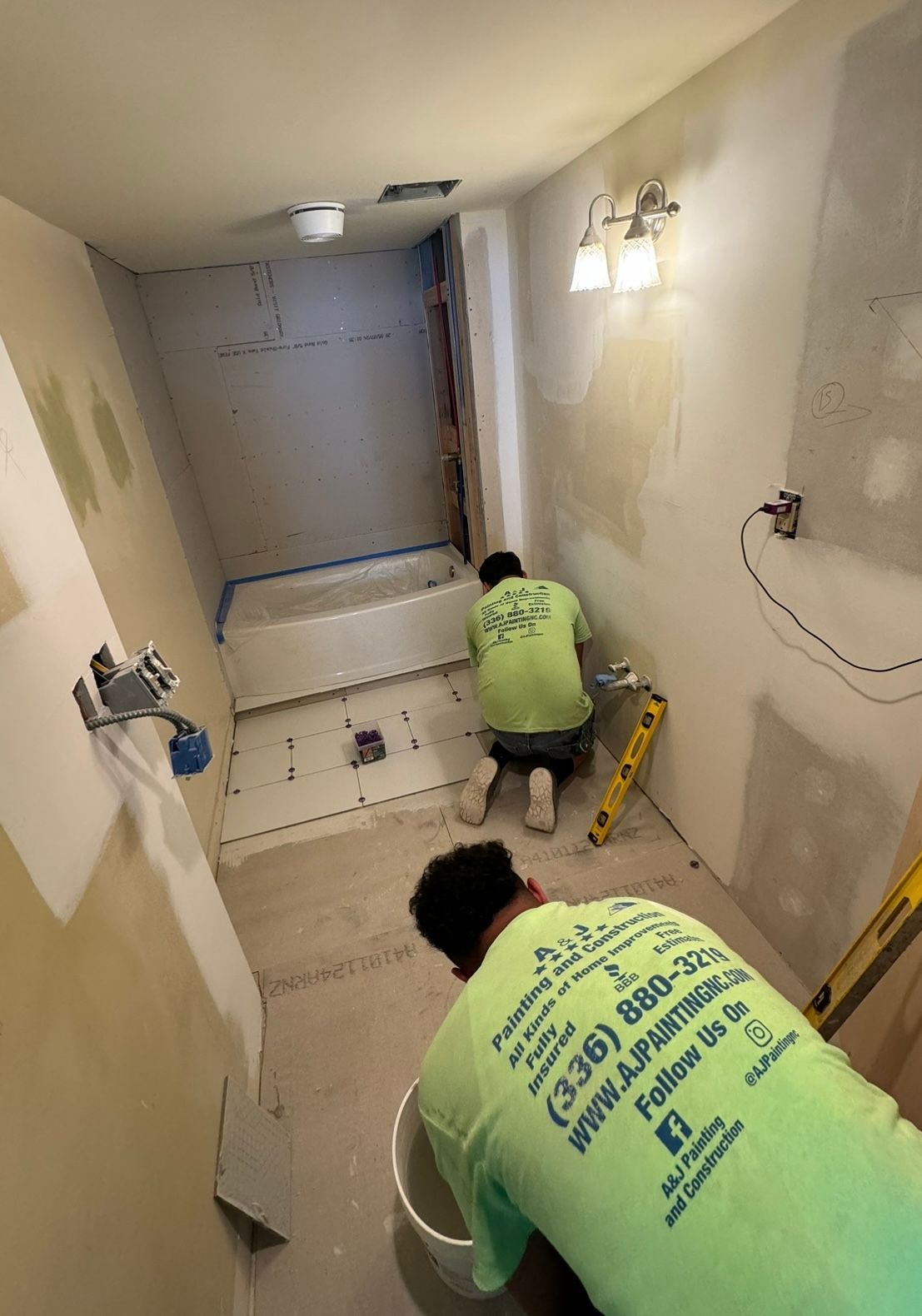 Two men in green shirts are working in a bathroom.