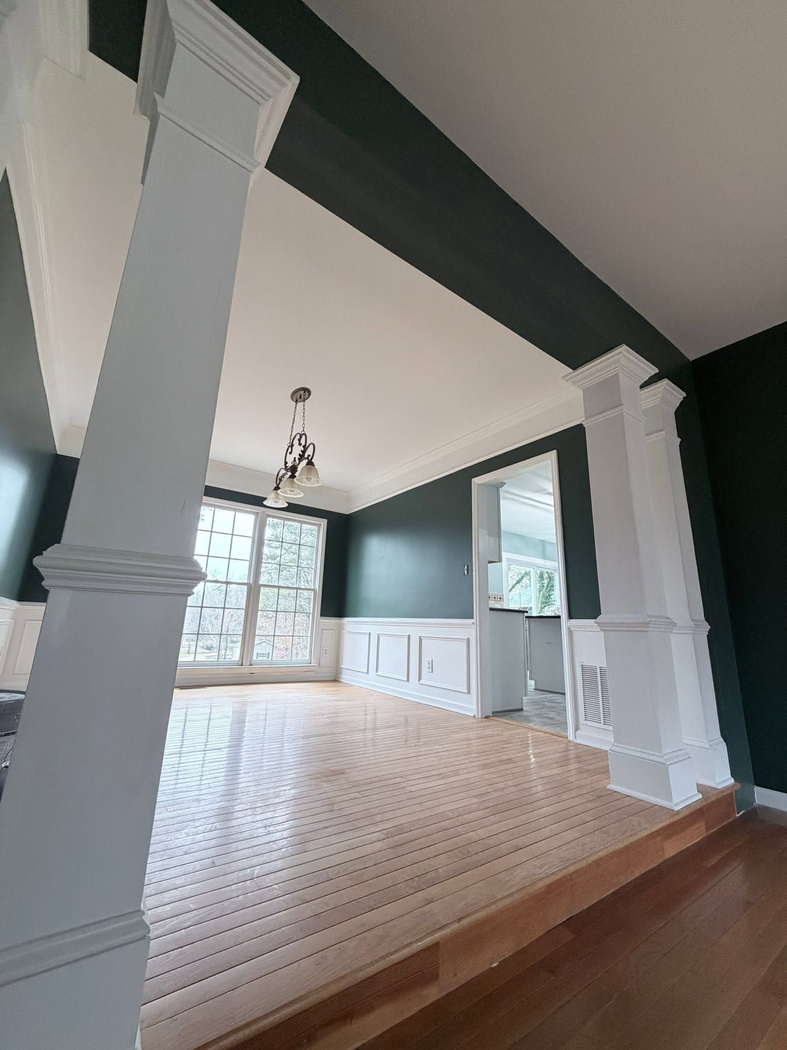 Empty dining room with white columns, patterned rug, dark green walls, and a chandelier near a large window