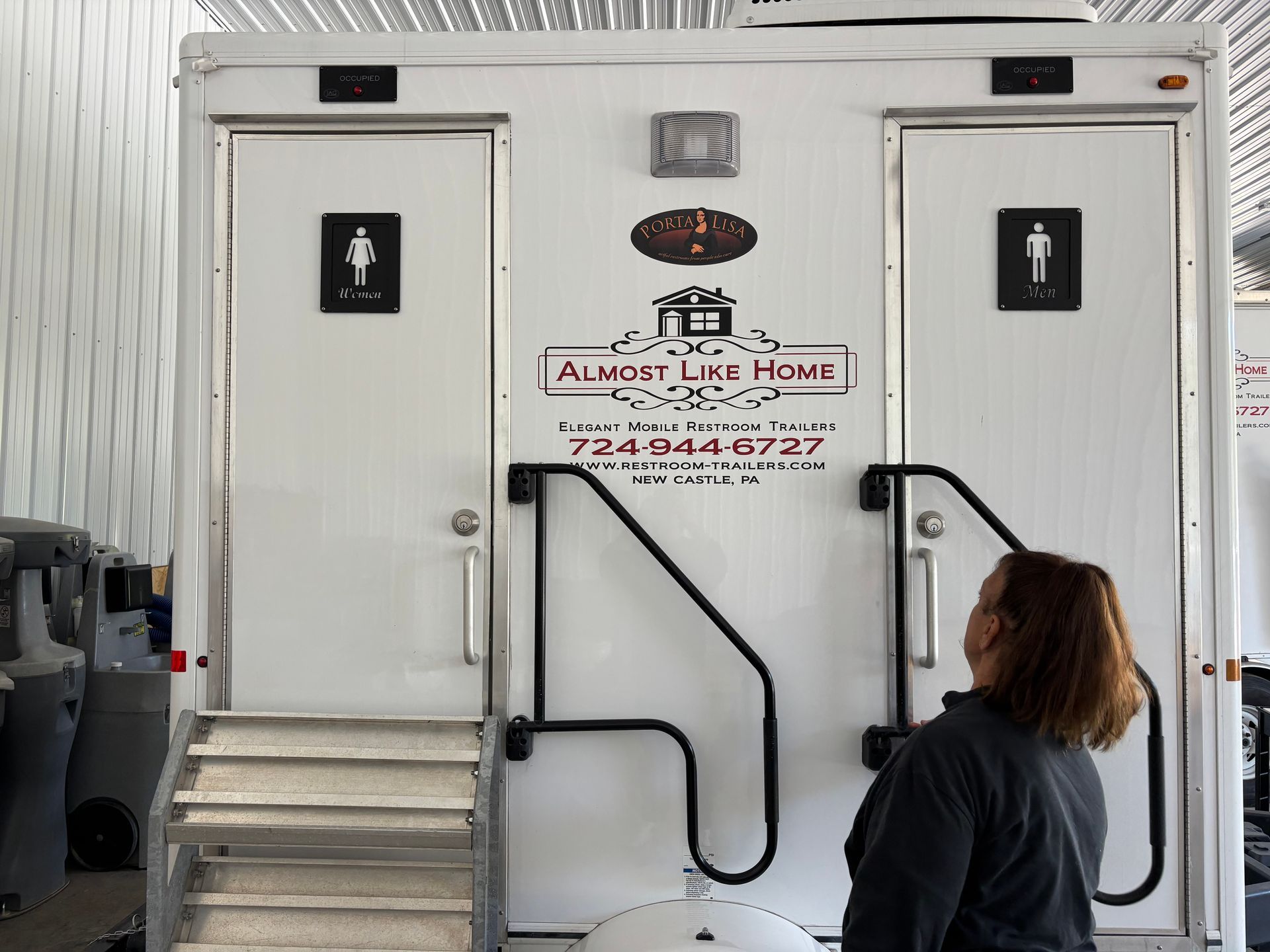 White portable restroom trailer with stairs and doors, with a person standing at the entrance.