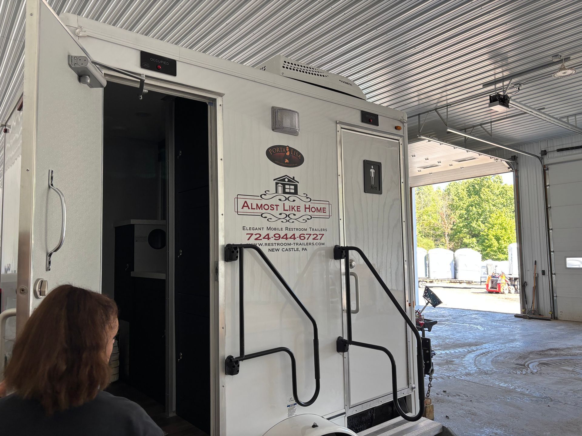 White ADA restroom trailer with open door, black handrails, under a covered outdoor area.