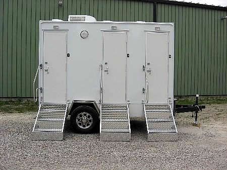 White portable restroom trailer with three doors and metal steps on gravel outside a green building.