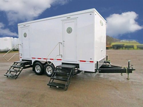 White portable trailer restroom with two black steps, parked outdoors on pavement under a cloudy sky