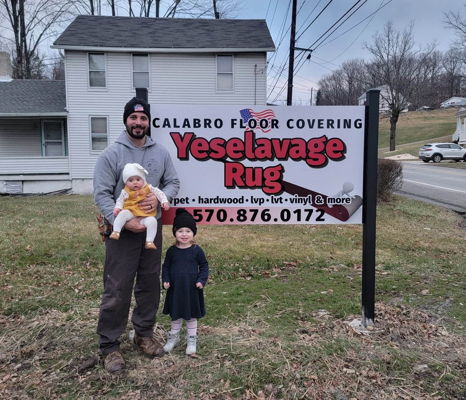 A man holding a baby stands in front of a yeselavage rug sign