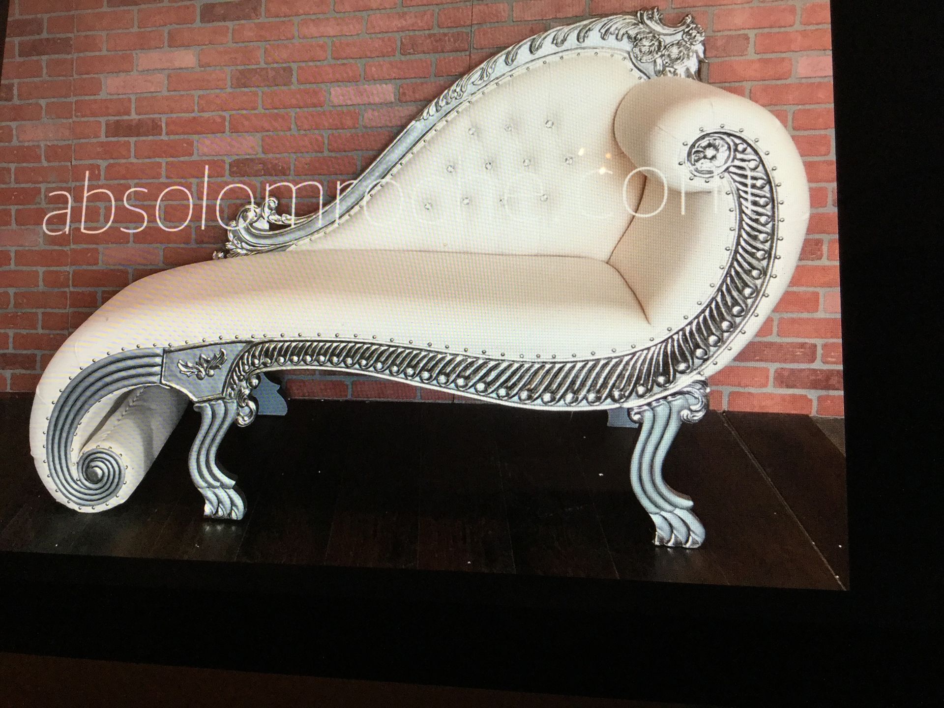 Cream tufted chaise lounge with ornate silver trim, shown against a brick wall.