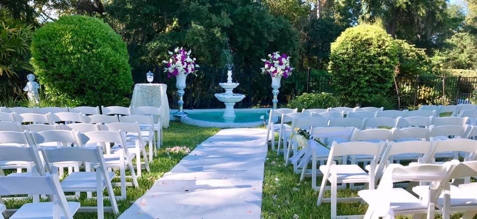 Outdoor wedding ceremony setup with white chairs lining a central aisle and a fountain in the garden backdrop