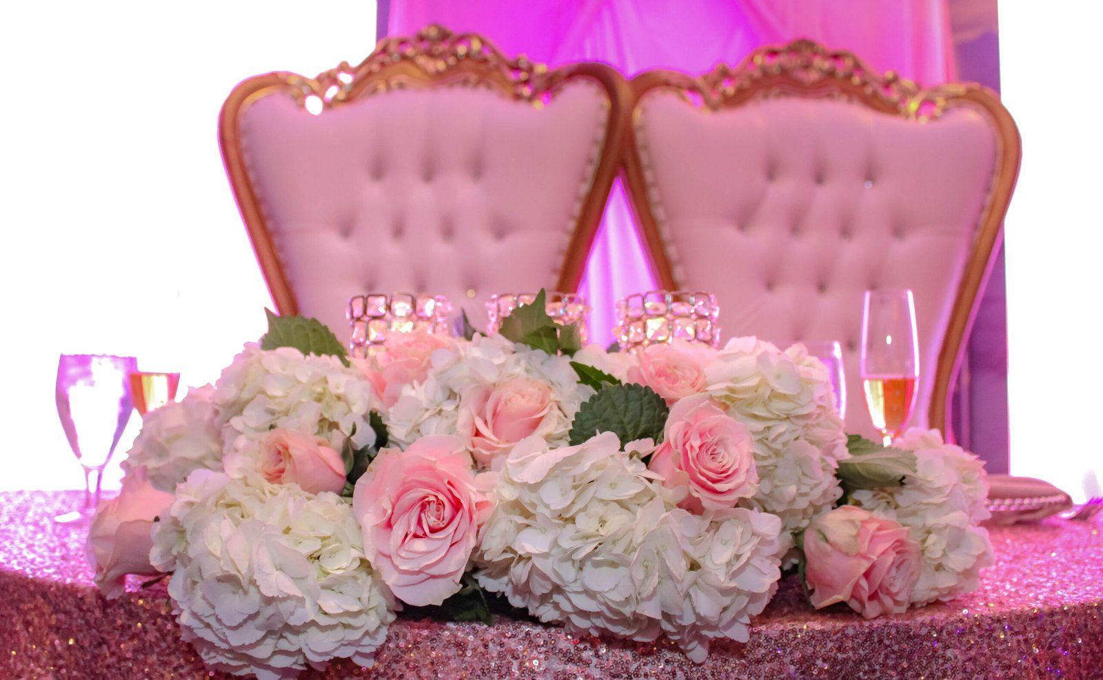Pink tufted chairs behind a pink glitter table with white and pink rose bouquets and wine glasses