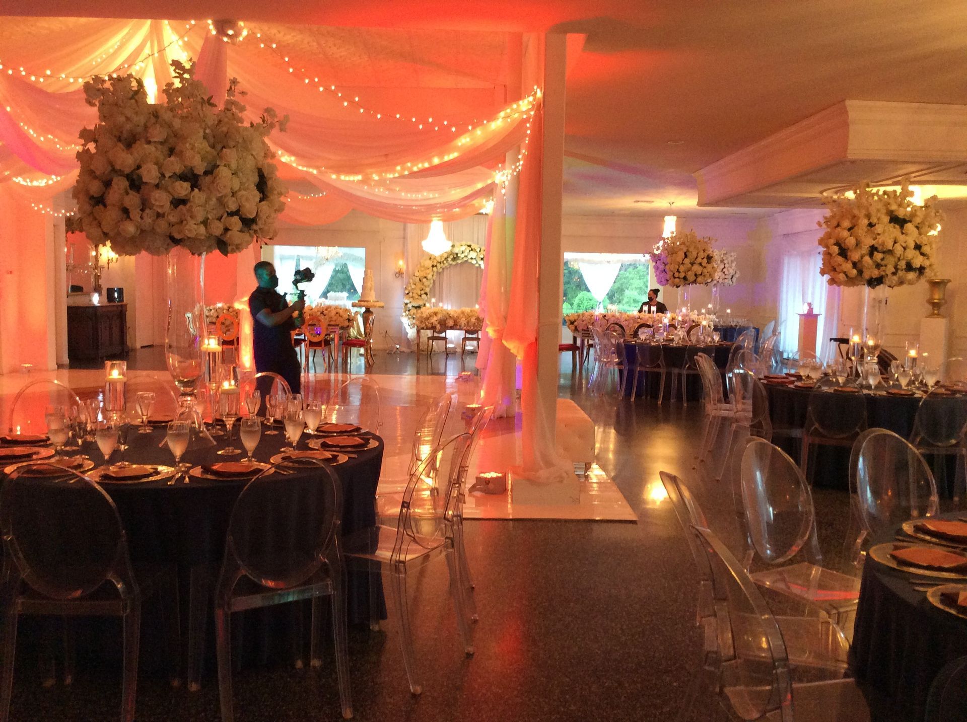 Elegant banquet hall with warm orange lighting, draped fabric, round tables, and floral centerpieces