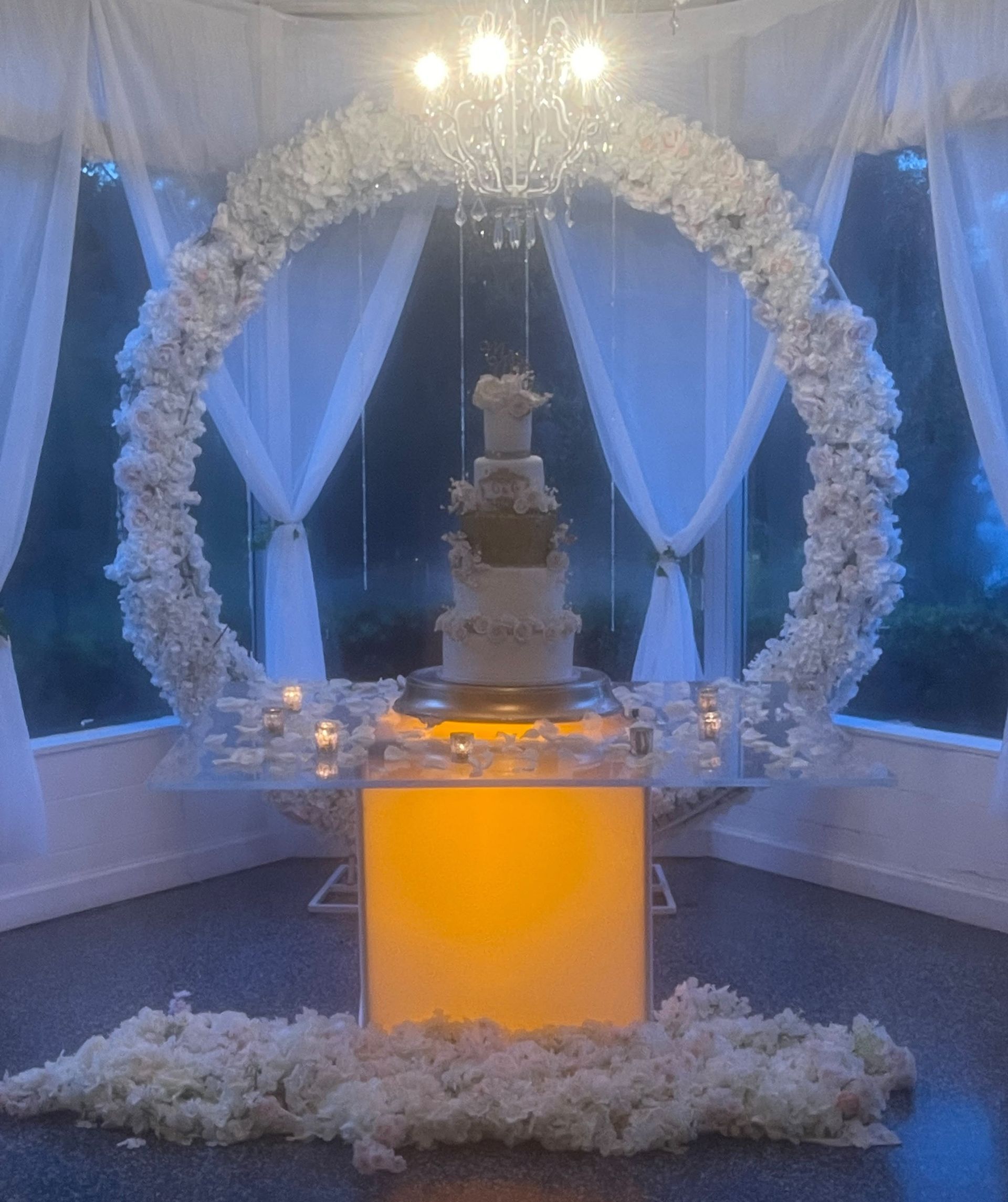 White floral wedding display with a tiered cake on an illuminated pedestal under a flower arch