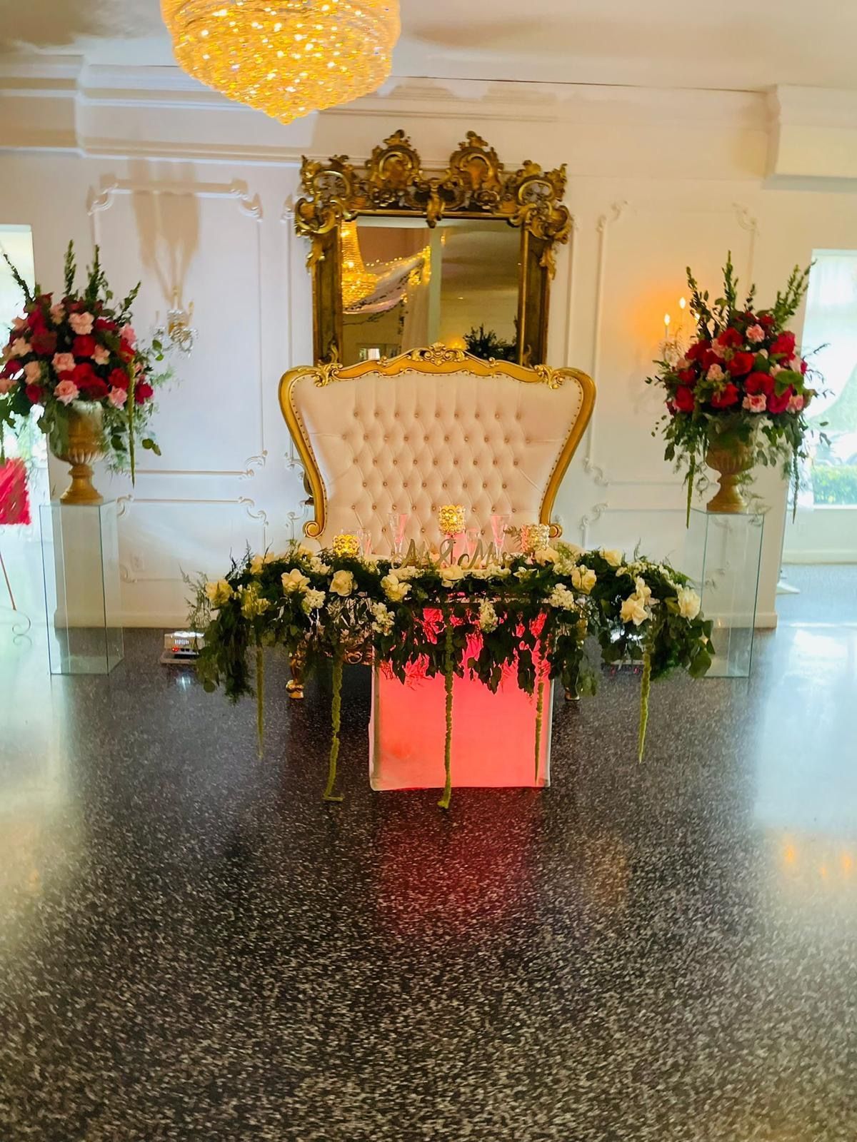 Elegant event stage with ornate gold chair, floral arrangements, and mirrored backdrop in a white ballroom